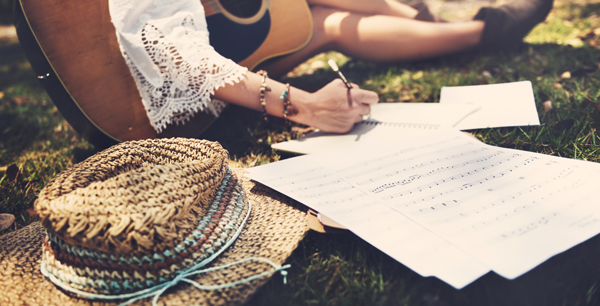 portion of a woman's body, holding a guitar in one hand and writing lyrics with the other as she sits in a park.