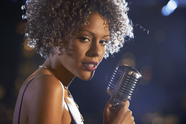 Black female with full hair and evening gown sings into mic.